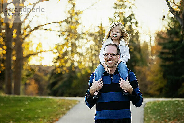Ein Vater trägt seine kleine Tochter auf den Schultern  während er im Herbst in einem Stadtpark spazieren geht; St. Albert  Alberta  Kanada