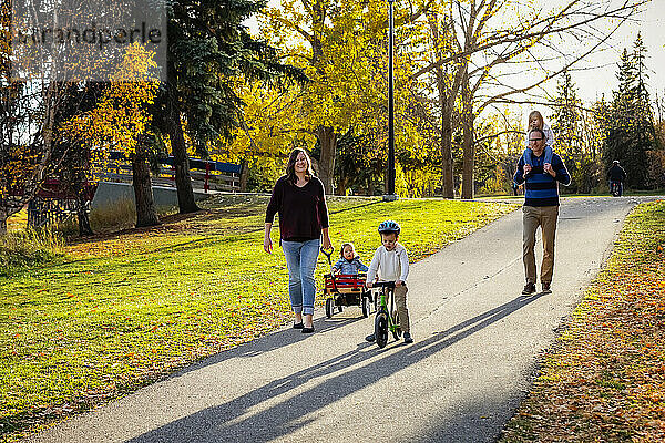 Eine Mutter zieht ihre Tochter mit Down-Syndrom in einem Wagen  der Vater hat die Tochter auf den Schultern und der Sohn fährt mit dem Fahrrad in einem Stadtpark während eines Familienausflugs in der Herbstzeit; St. Albert  Alberta  Kanada