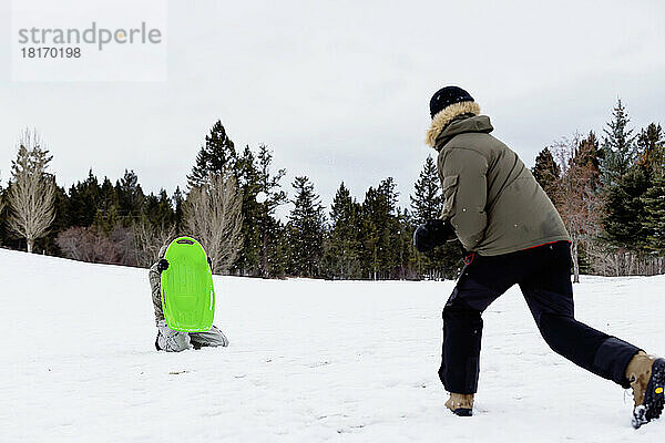 Ein Mann wirft Schneebälle auf einen anderen Erwachsenen  der einen Schlitten hält  um sie in einem Bergferienort zu blockieren; Fairmont Hot Springs  British Columbia  Kanada