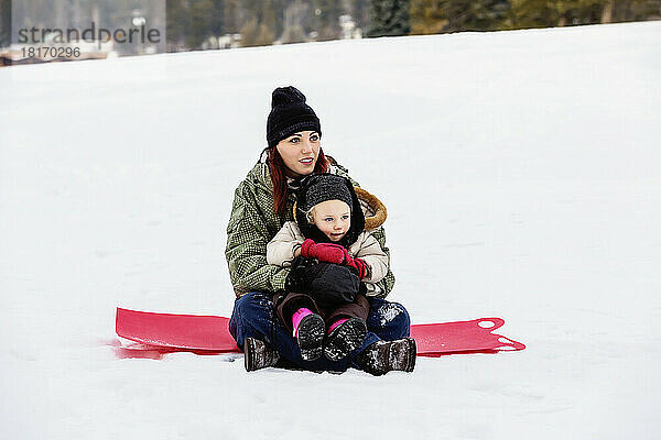 Mutter und junge Tochter sitzen auf einem Schlitten auf einem verschneiten Hügel in einem Bergferienort; Fairmont Hot Springs  British Columbia  Kanada