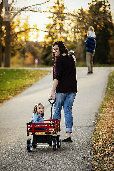 Eine Mutter zieht ihr Baby mit Down-Syndrom in einem Wagen entlang eines Flusses in einem Stadtpark mit ihrer Familie während der Herbstsaison; St. Albert  Alberta  Kanada
