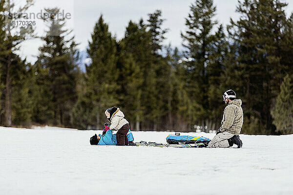 Ein Vater beobachtet zwei kleine Kinder  die beim Schlittenfahren im Schnee spielen; Fairmont Hot Springs  British Columbia  Kanada