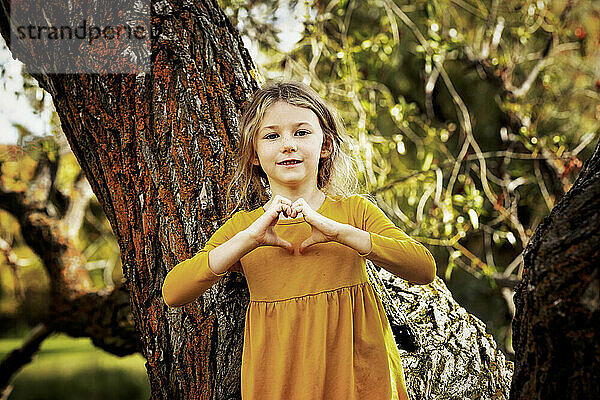 Junges Mädchen  das an einem warmen Herbstnachmittag in einem Stadtpark in der Nähe eines großen Baumes spielt und für ein Foto innehält  während sie mit ihren Händen eine Herzform der Liebe bildet; St. Albert  Alberta  Kanada