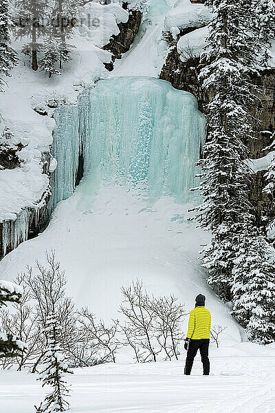 Frau beim Schneeschuhwandern auf einem schneebedeckten Weg mit einem Eisfall auf einer Klippe mit schneebedeckten Bäumen im Banff National Park; Lake Louise  Alberta  Kanada