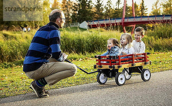 Ein Vater zieht seine kleinen Kinder in einem Wagen entlang eines Flusses in einem Stadtpark mit einer Brücke im Hintergrund während der Herbstsaison; sein kleines Mädchen hat das Down-Syndrom; St. Albert  Alberta  Kanada