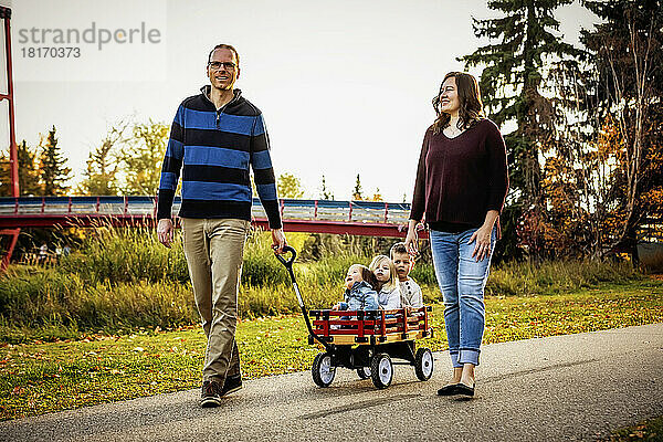 Ein Vater und eine Mutter ziehen ihre kleinen Kinder in einem Wagen in einem Stadtpark während der Herbstsaison  und ihr kleines Mädchen hat das Down-Syndrom; St. Albert  Alberta  Kanada