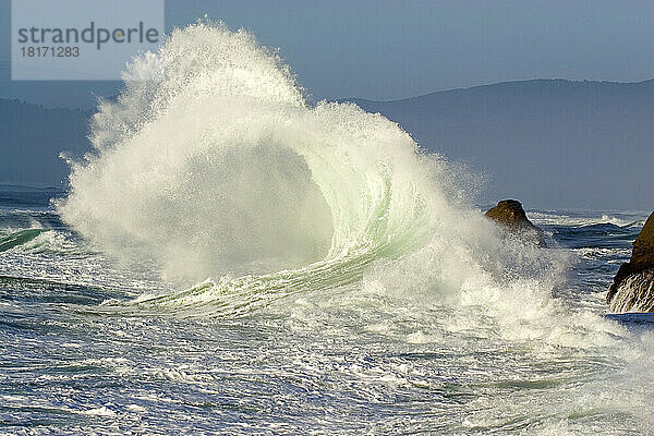Dramatische  hoch aufspritzende Welle  die sich an der Küste bricht  mit der Küstenlinie von Oregon im Hintergrund am Cape Kiwanda; Pacific City  Oregon  Vereinigte Staaten von Amerika