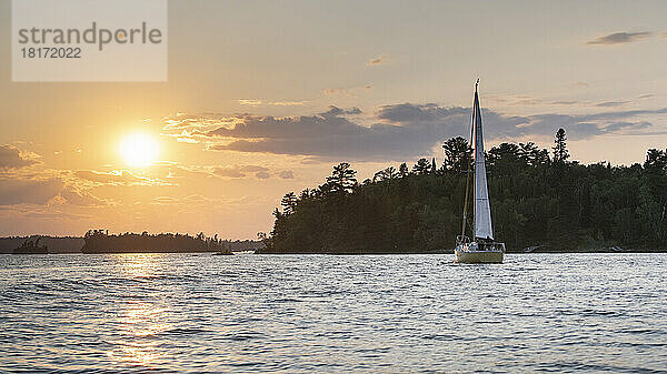 Segeln auf einem See bei Sonnenuntergang mit einer goldenen Sonne  die über der Silhouette der Landschaft und dem Wasser leuchtet  Lake of the Woods  Ontario; Kenora  Ontario  Kanada