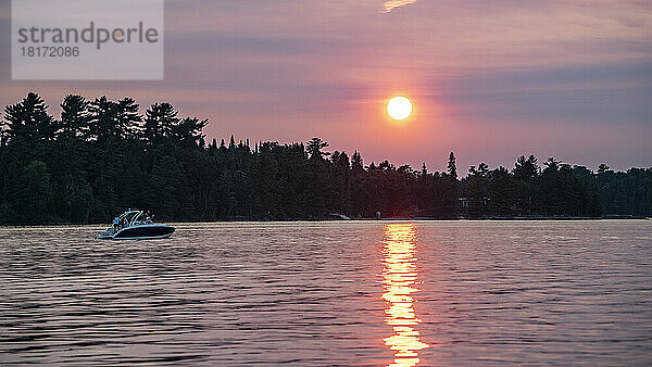 Bootfahren auf einem See bei Sonnenuntergang mit einer leuchtend rosa Sonne  die über der Silhouette der Landschaft und dem Wasser leuchtet  Lake of the Woods  Ontario; Kenora  Ontario  Kanada