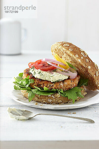 Lachsburger auf einem Vollkornbrötchen mit Salat  roten Zwiebeln  roten Tomaten  gelben Tomaten  Soße und frischem Dill auf einer weißen Tischplatte in einem High-Key-Ambiente