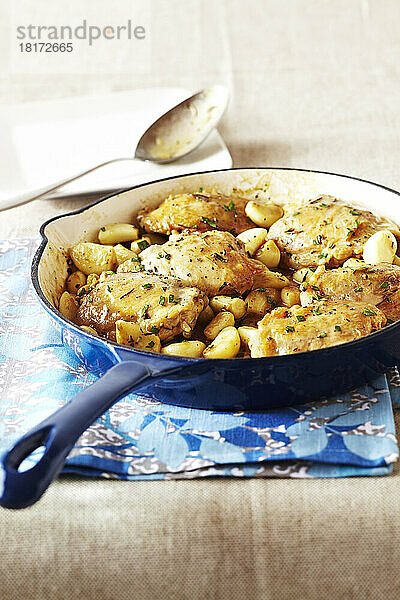 Blaue Bratpfanne mit Hähnchenschenkeln und Knoblauch auf einem gemusterten Geschirrtuch