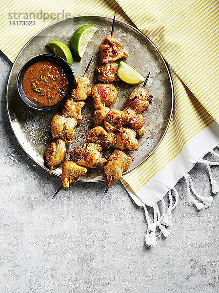 Hähnchen-Satay-Spieße mit Dip-Sauce und Limettenspalten auf einem Metalltablett auf gestreiftem Stoff