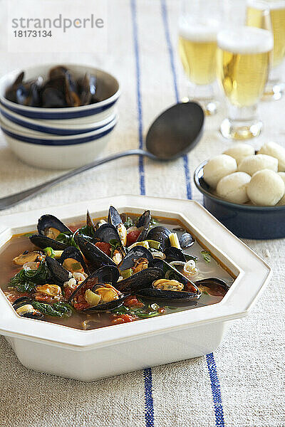 Servierschüssel Muschelsuppe mit Brötchen und Bier auf einer blau gestreiften Leinentischdecke