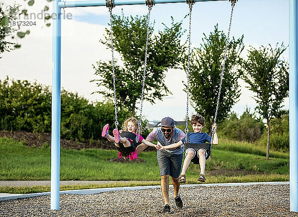 A father pushes a young girl and boy on swings on a swing set at a playground; St. Albert  Alberta  Canada