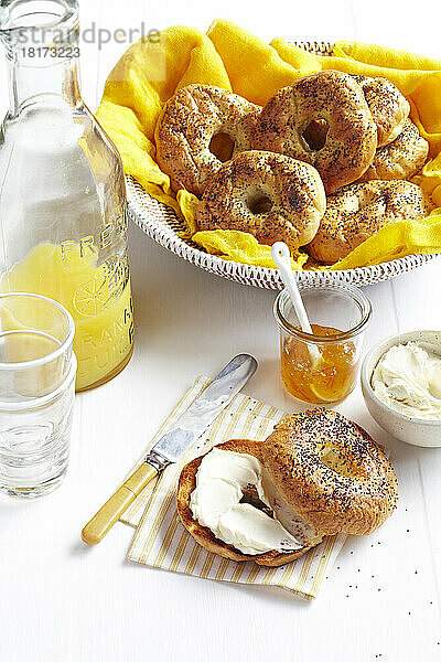 Bagel mit Frischkäse und Orangenkonfitüre  serviert mit frischem Orangensaft und einem Korb voller Bagels im Hintergrund.