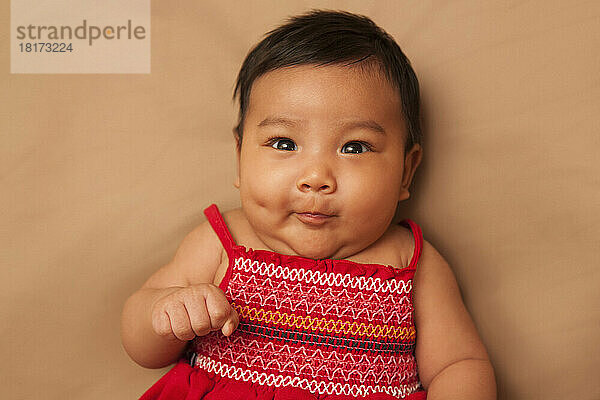 Nahaufnahme eines asiatischen Babys  das auf dem Rücken liegt  ein rotes Kleid trägt  in die Kamera blickt und ein lustiges Gesicht macht  Studioaufnahme auf braunem Hintergrund