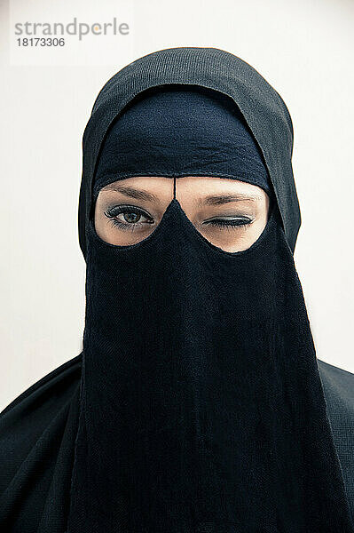 Nahaufnahme einer jungen Frau in schwarzem  muslimischem Hijab und muslimischer Kleidung  die zwinkert und in die Kamera blickt  Augen mit Augen-Make-up  Studioaufnahme auf weißem Hintergrund