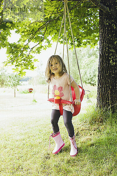 3-jähriges Mädchen in Gummistiefeln sitzt in roter Schaukel im Hinterhof  blickt in die Kamera und lächelt  Schweden