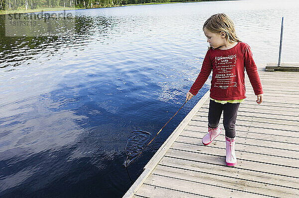 3-jähriges Mädchen in rotem Hemd auf einem Pier  einen Stock in der Hand und im Wasser spielend  Schweden