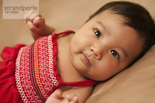 Close-up portrait of Asian baby lying on back  wearing red dress  looking at camera  studio shot on brown background