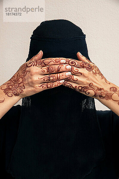 Nahaufnahmeporträt einer Frau mit schwarzem muslimischem Hijab und muslimischer Kleidung  die Hände bedecken die Augen und zeigen mit Henna im arabischen Stil bemalte Hände  Studioaufnahme auf weißem Hintergrund