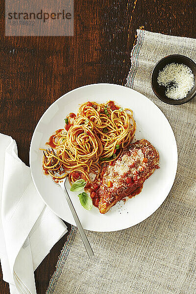 Teller mit Hühnerparmesan mit Spaghetti und Tomatensauce und einer Beilage Käse mit Gabel und Serviette auf einem Holzhintergrund