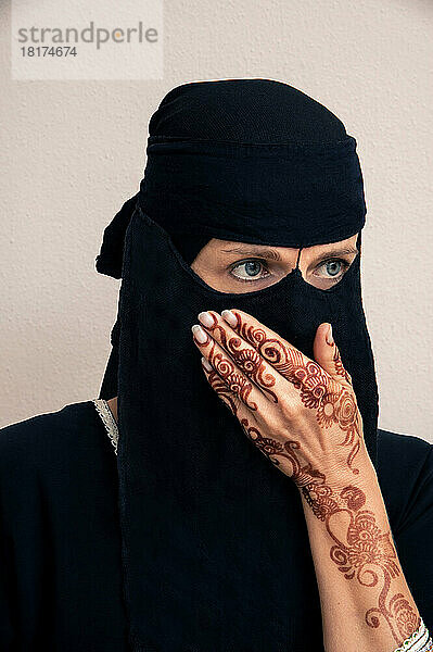 Nahaufnahme einer Frau im schwarzen muslimischen Hijab und in muslimischer Kleidung  die zur Seite schaut  die Hand vor dem Mund hält und mit Henna im arabischen Stil bemalte Arme und Hände zeigt  Studioaufnahme auf weißem Hintergrund