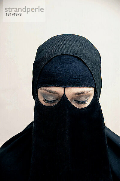 Nahaufnahme einer jungen Frau in schwarzem  muslimischem Hijab und muslimischer Kleidung  mit geschlossenen Augen und Augen-Make-up  Studioaufnahme auf weißem Hintergrund