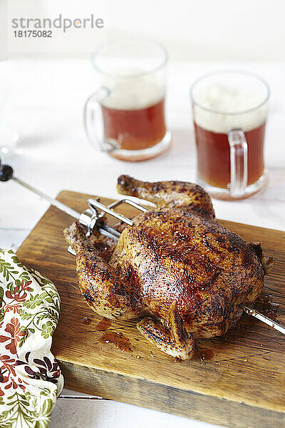 Ganzes Hähnchen auf einem Drehspieß  auf einem Schneidebrett platziert  mit zwei Krügen Bier