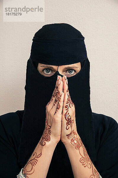 Nahaufnahme einer Frau mit schwarzem Hijab und muslimischer Kleidung  die in die Kamera blickt  mit betenden Händen und mit Henna im arabischen Stil bemalten Armen und Händen  Studioaufnahme auf weißem Hintergrund