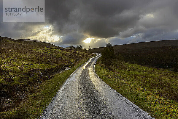Kurvenreiche Straße im Hochland mit Sonne  die durch die dunklen Wolken bei Glen Coe in Schottland  Vereinigtes Königreich scheint