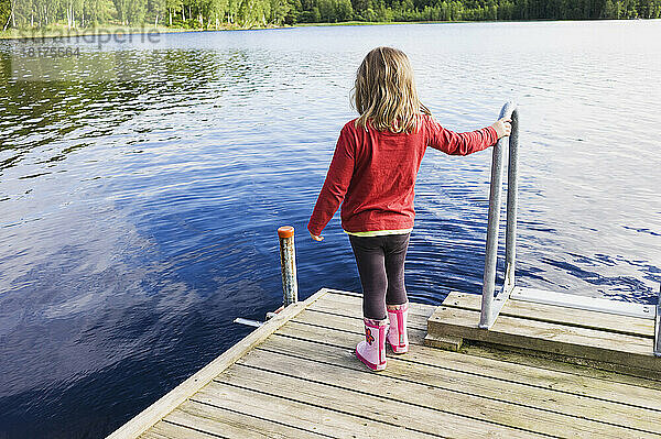 3 Jahre altes Mädchen im roten Hemd auf einem Holzsteg mit Blick auf einen See  Schweden