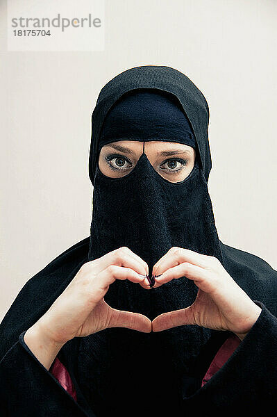 Nahaufnahme einer jungen Frau in schwarzem  muslimischem Hijab und muslimischer Kleidung  die mit den Händen ein Herz formt  in die Kamera blickt  Augen mit Augen-Make-up zeigt  Studioaufnahme auf weißem Hintergrund