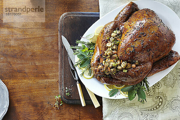 Holiday whole cooked turkey with glaze  herbs  and stuffing on a bed of fresh herbs and lemons. On a white serving platter with carving utensils on a wooden tabletop.