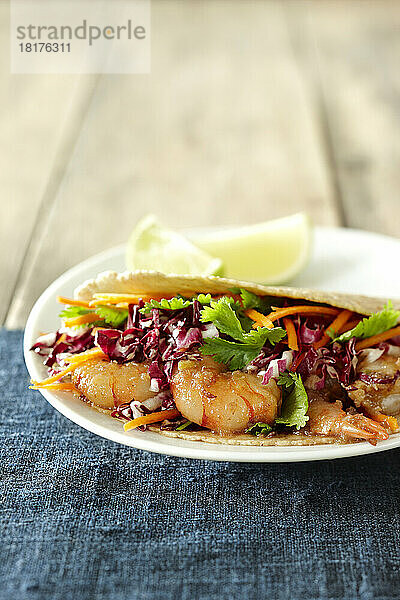 Einzelportion Garnelen-Taco mit Rotkohl  Karotte und Koriander auf Maistortillas mit Limettenscheiben auf Holzhintergrund