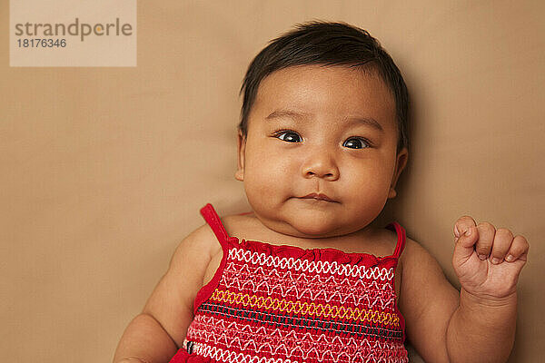 Close-up portrait of Asian baby lying on back  wearing red dress  looking at camera and smiling  studio shot on brown background