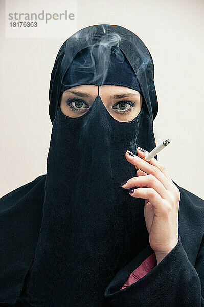 Nahaufnahme einer jungen Frau in schwarzem  muslimischem Hijab und muslimischer Kleidung  die eine Zigarette in der Hand hält und raucht  in die Kamera blickt  Augen mit Augen-Make-up zeigt  Studioaufnahme auf weißem Hintergrund