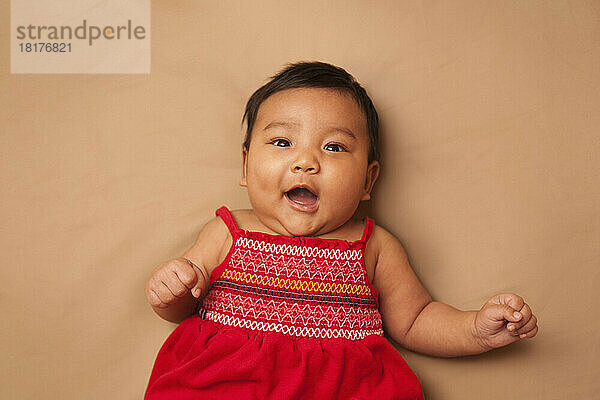 Close-up portrait of Asian baby lying on back  wearing red dress  looking at camera and smiling  studio shot on brown background