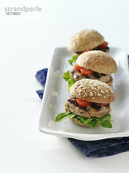 Drei gegrillte Puten-Burger-Slider auf Vollkornbrötchen mit Tomaten und Salat auf einer weißen Platte in einer hohen Tischdekoration mit einer blauen Stoffserviette.