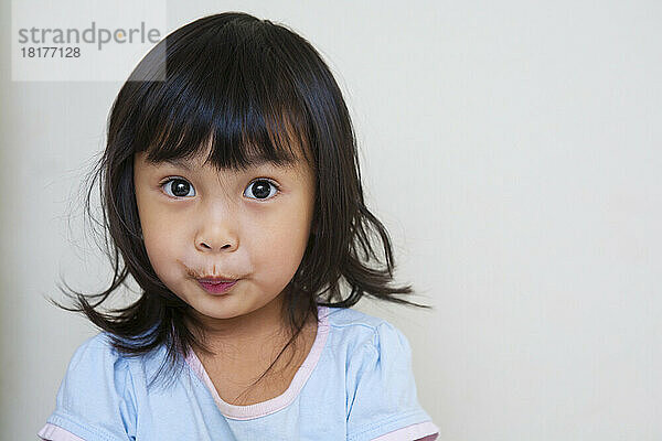 Nahaufnahmeporträt eines asiatischen Kleinkindmädchens  das mit überraschtem Gesichtsausdruck in die Kamera blickt  Studioaufnahme auf weißem Hintergrund