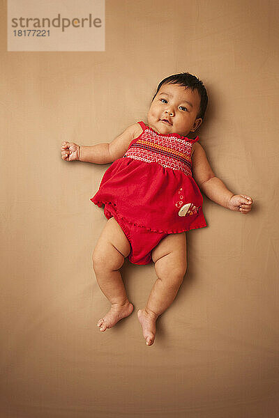 Porträt eines asiatischen Babys  das auf dem Rücken liegt  ein rotes Kleid trägt  in die Kamera blickt und lächelt  Studioaufnahme auf braunem Hintergrund