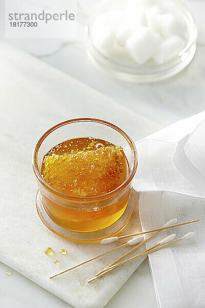Ein kleines Glasgefäß mit klarem  goldenem Honig und eine Honigwabe mit Wattestäbchen und Wattebällchen. Hohes Schlüsselimage von Honig und Schönheitsprodukten.