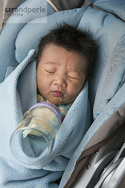 Close-up of swaddled two week old  newborn Asian baby girl  lying in car seat  disgusted with her milk bottle