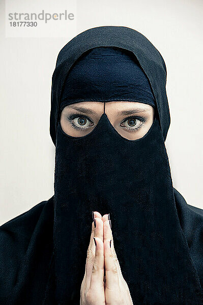 Nahaufnahmeporträt einer jungen Frau in schwarzem  muslimischem Hijab und muslimischer Kleidung  mit betenden Händen  Blick in die Kamera  Augen mit Augen-Make-up  Studioaufnahme auf weißem Hintergrund