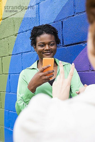Lächelnde Frau hält Smartphone in der Hand und blickt Freundin an  die an einer bunten Wand ein Friedenszeichen zeigt