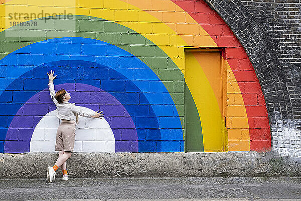 Junge Frau mit erhobener Hand stützt sich auf eine an die Wand gemalte Regenbogenfahne