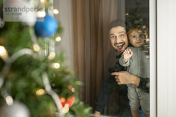 Glücklicher Vater und Sohn  die durch das Fenster auf den Weihnachtsbaum schauen