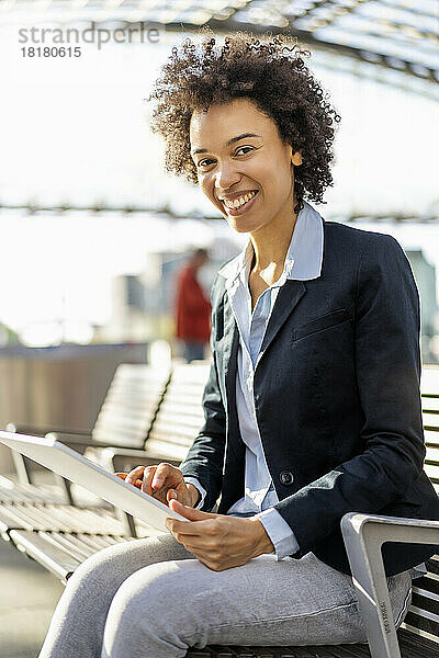 Glückliche Geschäftsfrau mit Tablet-PC sitzt auf einer Bank am Bahnhof