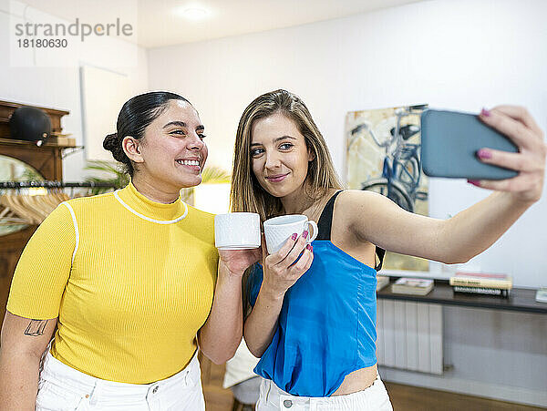 Lächelnde Frau hält eine Kaffeetasse in der Hand und macht mit einem Freund ein Selfie über ihr Mobiltelefon
