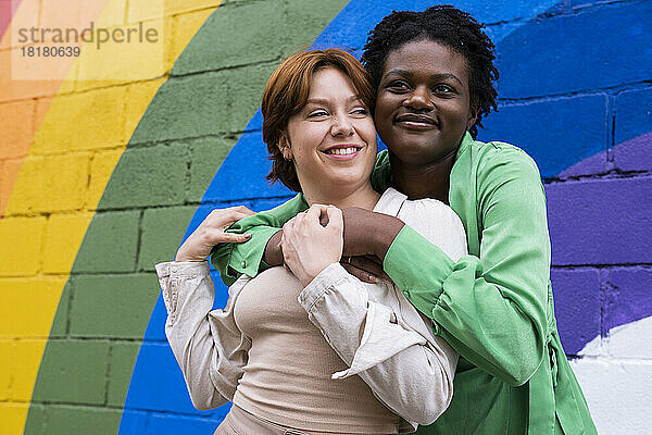 Lächelnde Frau umarmt ihre Freundin vor einer an die Wand gemalten Regenbogenfahne